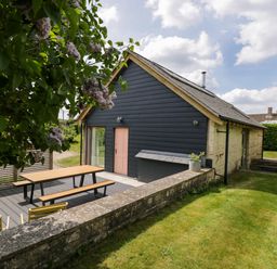 A house with dark siding and a pink door with a picnic table on a deck and grass surrounding the area at The Barn at Daubeneys in Colerne
