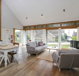 A living room with two gray sofas a white round coffee table and a dining table with chairs near large windows at The Barn at Daubeneys in Colerne