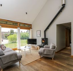 A living room with large windows a sofa armchair coffee table wood stove and television at The Barn at Daubeneys Colerne
