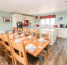 A dining area with a wooden table set for ten and a kitchen with white cabinets and appliances at Harmon Vale in Solva