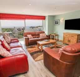 A living room with three leather sofas a wooden coffee table a flat screen TV on the wall large windows with red blinds and a view of houses and the sea at Harmon Vale in Solva