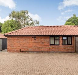 A red brick building with a tiled roof and three windows facing a paved open area with trees in the background at The Nest Box in Diss