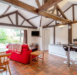 A living area with a red sofa a wooden round table wooden chairs a small wooden side table a wall-mounted TV and a black breakfast bar with two stools at The Nest Box in Diss