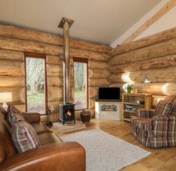 A living room with a leather sofa, plaid armchair, wood burning stove, TV on a stand, wooden shelves, and large windows at Moorhen Lodge in North Kessock