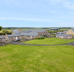 A group of stone buildings with slate roofs surrounded by green fields and paved paths at St  Cruz in Embleton