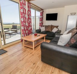 A living room with black sofas and cushions a wooden coffee table a wall mounted tv and glass doors leading to a patio with outdoor furniture at Newton View in Embleton