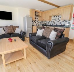 A living room with two black sofas and a wooden table in front of a kitchen with wooden cabinets and a silver refrigerator at Newton View in Embleton