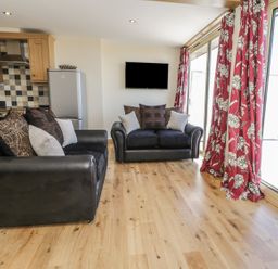 An open living area with two black sofas with cushions a wall-mounted television wooden kitchen cabinets and large sliding glass doors with red floral curtains at Mill View in Embleton