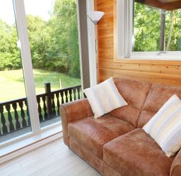 A living room with a brown sofa with pillows next to large windows showing a grassy yard at Kipling Lodge in Rudyard
