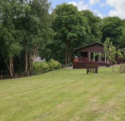 A wooden house with sliding glass doors surrounded by trees and a large grassy lawn at Kipling Lodge in Rudyard