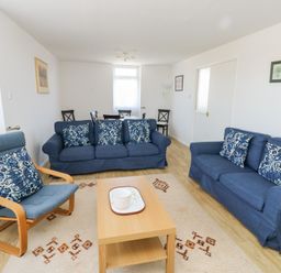 A living room with two blue sofas and one blue armchair with patterned cushions a wooden coffee table and a dining table with chairs at La Capsule in Dawlish Warren