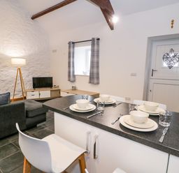 A kitchen and living area with a dining counter set for four people a gray sofa a TV on a wooden stand a floor lamp and a white door with a heart decoration at Cornel Glyd in Talybont near Barmouth