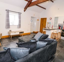 A living room with gray cushions on a sofa and a kitchen area with stools and a stove at Cornel Glyd in Talybont near Barmouth