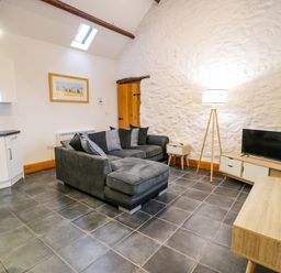 A living room with a grey sectional sofa a floor lamp a wooden media cabinet with a television and a kitchen area at Cornel Glyd in Talybont near Barmouth