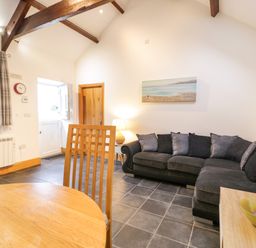 A living room with a round wooden dining table wooden chair a dark gray sectional sofa wall art and a door at Sisial-y-Mor in Talybont near Barmouth