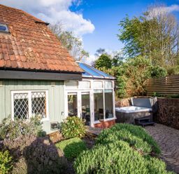 A garden area with bushes a patio with a hot tub next to a conservatory and a house with tile roof and green walls at Fig Tree Cottage in Bickleigh