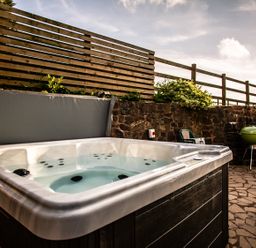 An outdoor hot tub on a stone patio with a wooden fence stone wall green barbecue and plastic chairs at Fig Tree Cottage in Bickleigh