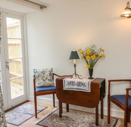 A small seating area with two wooden chairs with floral cushions a small wooden table with a lamp and flowers beside a glass door with floral curtains at Tick Tock Cottage in Marnhull