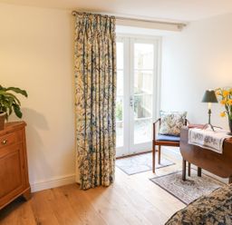 A room with wooden cabinet, floral curtains, wooden chair with cushion, small table with lamp and flowers at Tick Tock Cottage in Marnhull