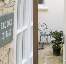 A stone wall with a Tick Tock Cottage sign and an open glass door leading to a small outdoor area with a metal chair and potted plants at Tick Tock Cottage in Marnhull