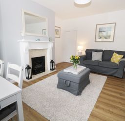 A living room with a gray sofa, a gray ottoman with flowers on it, a white fireplace with two black lanterns, a white dining table with chairs, and framed pictures on the walls at Rose Cottage in Lesbury near Alnmouth