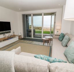 A living room with a sofa with cushions a television and a sliding glass door showing an outdoor view at Chapel Rock View The Dunes in Perranporth