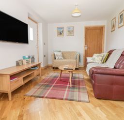 A living room with a brown leather sofa, a patterned armchair, a wooden coffee table, a plaid rug, a wooden TV stand, and a wall-mounted television at The Stable at Church Farm House Limpenhoe near Reedham