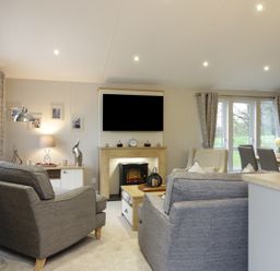 A living room with gray armchairs a TV above a fireplace and a dining area with a table at Woodstock Lodge in Wemyss Bay