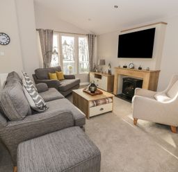A living room with grey sofas, a wooden coffee table, a fireplace, a wall-mounted television, and large windows at Woodstock Lodge in Wemyss Bay