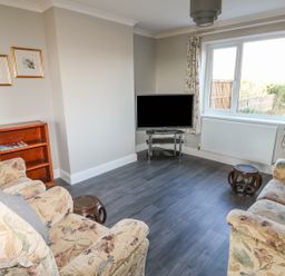 A living room with two floral armchairs a small wooden stool a TV on a glass stand two framed pictures on the wall and floral curtains at 27 Whitegates in Ludham