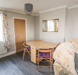 A dining area with a wooden table and four chairs with purple cushions near a floral sofa and a wooden door at 27 Whitegates in Ludham