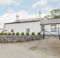 A white cottage with two chimneys a stone wall green bushes and a black metal gate at Pen Y Bryn Cottage in Llandudno