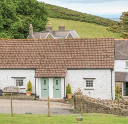 A white cottage with green doors and a tiled roof surrounded by grass and trees at Wren Cottage in Ilfracombe