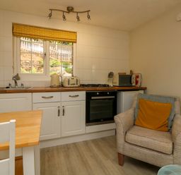 A kitchen with white cabinets a wooden countertop window with yellow blind and an armchair with cushions at Swallow Cottage in Ilfracombe