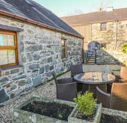 An outdoor patio area with a round glass table and four wicker chairs next to stone buildings with wooden windows at Beudy Bach in Y Felinheli