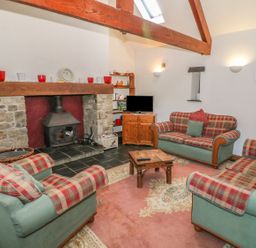 A living room with plaid armchairs and sofas around a wood stove and a television on a wooden cabinet at Beudy Bach in Y Felinheli