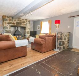 A living room with brown sofas facing a stone fireplace with a wood stove a red floor lamp wooden floor and a window with yellow curtains at Hendre in Y Felinheli