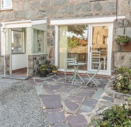 An outdoor patio area with stone flooring a small round table with two chairs potted plants and flowers and a stone building with a sliding glass door at Y Cilgwyn in Trawsfynydd