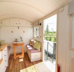 A small dining area with bench seating, table, cushions, and kitchen counter next to an open door in Shepherd's View in Rhayader