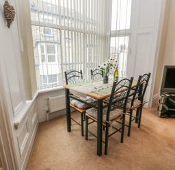 A dining area with a table set for four by large windows with vertical blinds and a television on a stand at Flat 4 in Whitby