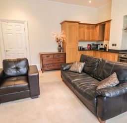 A living area with a black leather armchair and sofa with cushions next to a wooden kitchen and wooden chest of drawers at Flat 4 Whitby