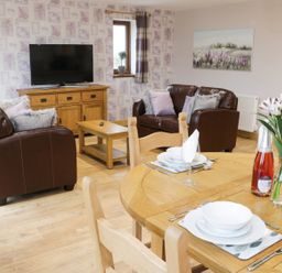 A living room with brown leather sofas a wooden coffee table a wooden TV stand with a television and a dining table set with white dishes and flowers at Gardener's Lodge in Nantwich