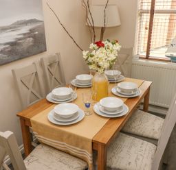 A dining table set for six with white plates bowls glasses and a flower vase in a room with six chairs a painting and a window with blinds at Sandy Bay House in Saundersfoot