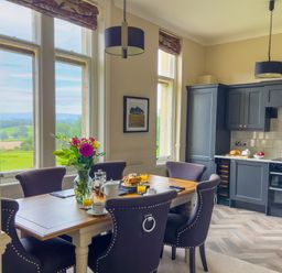 A kitchen and dining area with a wooden table set with breakfast items and windows showing countryside views at Geltsdale East Wing in Wetheral