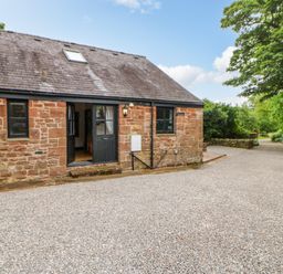 A stone house with black framed windows and a door opening to a gravel driveway surrounded by greenery at The Old Byre in Appleby-in-Westmorland
