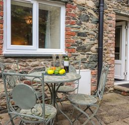 An outdoor patio area with a metal table and four chairs with a plate of fruit and a bottle of wine against a stone wall at Maple Cottage in Keswick