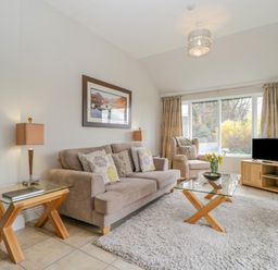 A living room with a beige sofa and armchair a glass coffee table with flowers and a wooden TV stand with a television at Garden Cottage in Keswick