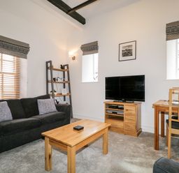 A living room with a black sofa, wooden coffee table, TV on wooden stand, shelving unit, and dining table at The Old Hayloft in Keswick