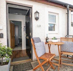 An outdoor seating area with a wooden table and two chairs with cushions and a plant near an open door at The Old Hayloft in Keswick