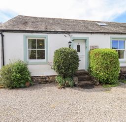 The front view of a white cottage with two windows a white door bushes and a wooden bench at Mell View Cottage in Ullswater